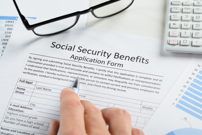 Close-up Of Hand With Pen And Eyeglasses Over Social Security Benefits Application Form