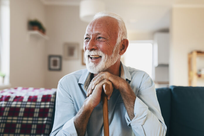 Portrait of happy senior man sitting at home with walking stock and smiling.Portrait of nice cheerful positive cheery stylish old man wearing checked shirt leaning on cane in white light modern interior studio room new house