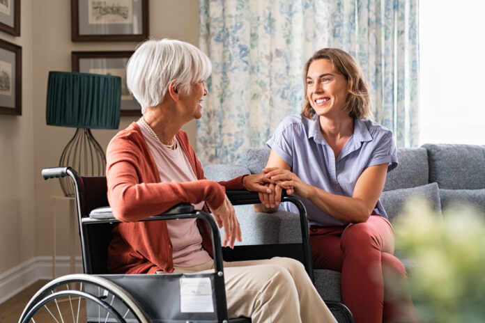 Mature woman comforting senior mom sitting on wheelchair at nursing home. Cheerful woman talking to old disabled mother in wheelchair at elder care centre. Loving caregiver taking care of elderly woman at home.