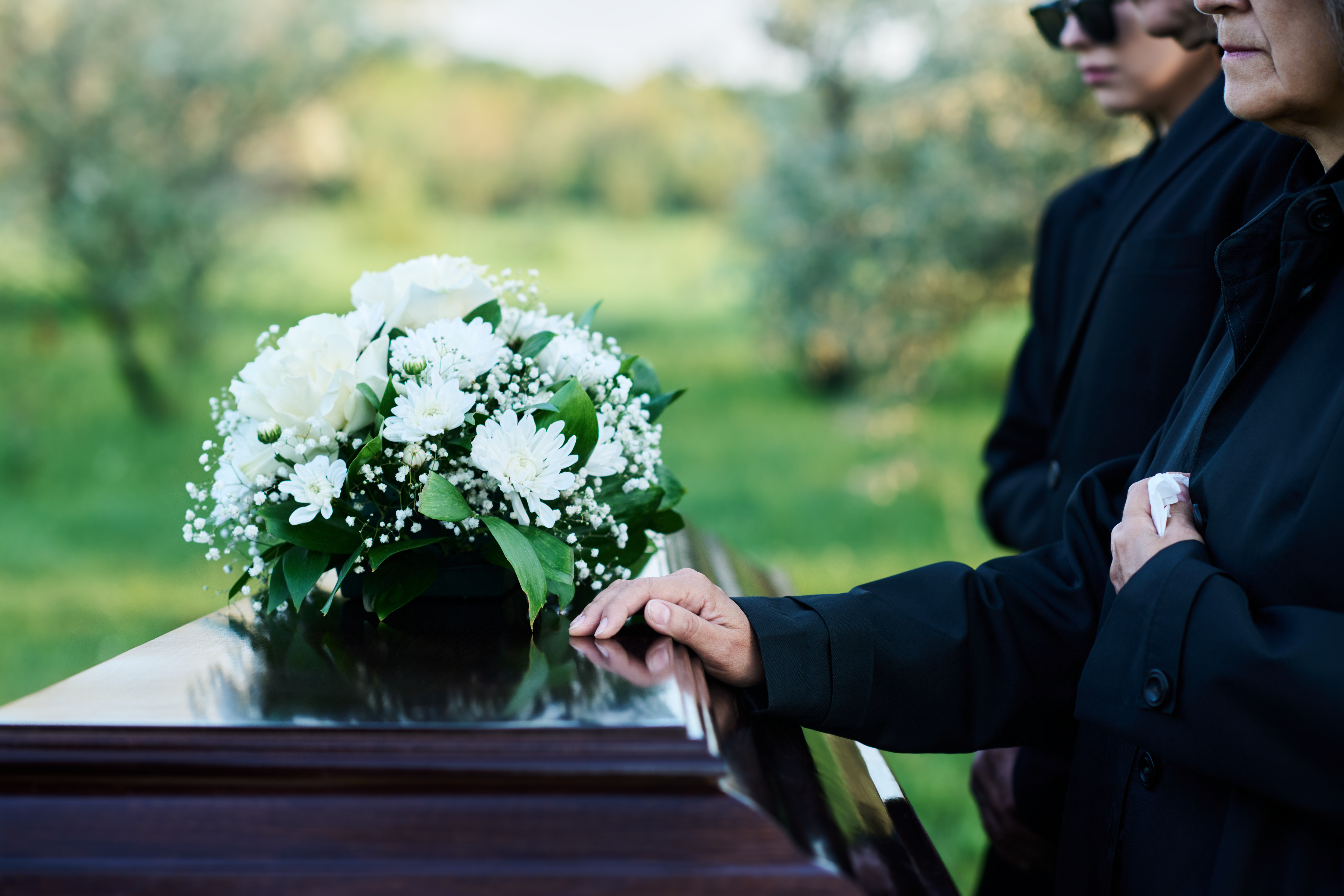 Cropped shot of mature woman and her family in mourning attire standing in front of coffin with closed lid with fresh white chrysanthemums