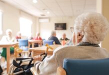 Back view of senior woman with short white hair sitting on chair with foldable walker with friends in blurred background at nursing home