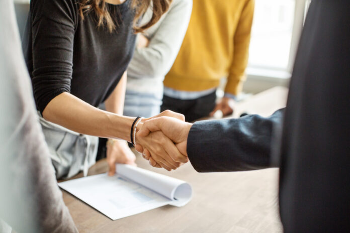 Two people shaking hands over a table with documents, symbolizing agreement or partnership, while others stand nearby in a casual office setting.