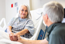A senior woman in the hospital room recovering, while her husband supports her.