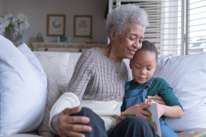 A beautiful senior woman sits on a couch with her granddaughter as they read a story together while smiling.